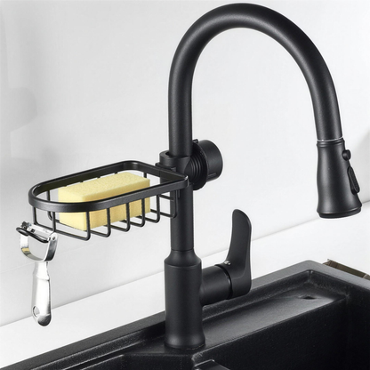 Black wire sink caddy clamped around the neck of a black pull-down kitchen faucet, holding a yellow sponge, with a chrome peeler hanging from the hook on its left end