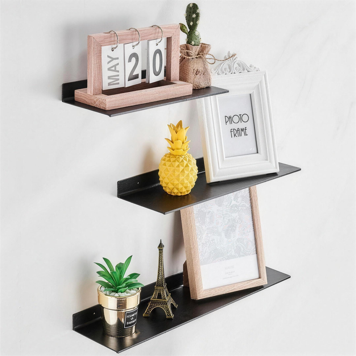 Three black floating shelves on a wall holding a desk calendar, photo frames and small decor