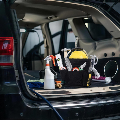 Black foldable cleaning caddy in an open car trunk holding a spray bottle, tape and tools, with cloths beside it